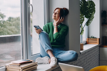 Thoughtful woman in headphones holding smart phone while sitting on the window sill at home