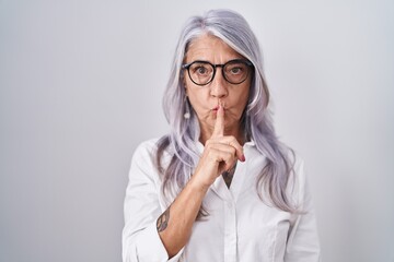 Middle age woman with tattoos wearing glasses standing over white background asking to be quiet with finger on lips. silence and secret concept.