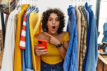 Young hispanic woman searching clothes on clothing rack using smartphone surprised pointing with finger to the side, open mouth amazed expression.