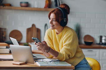Joyful young woman in headphones using technologies while sitting at the kitchen counter