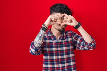 Young hispanic man with beard standing over red background doing heart shape with hand and fingers smiling looking through sign