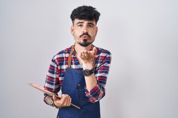 Young hispanic man with beard wearing waiter apron holding clipboard looking at the camera blowing a kiss with hand on air being lovely and sexy. love expression.