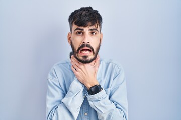 Young hispanic man with beard standing over blue background shouting suffocate because painful strangle. health problem. asphyxiate and suicide concept.