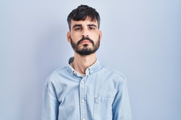 Young hispanic man with beard standing over blue background relaxed with serious expression on face. simple and natural looking at the camera.