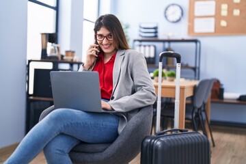 Young hispanic woman business worker using laptop talking on the smartphone at office