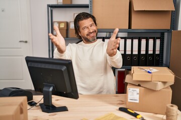 Handsome middle age man working at small business ecommerce looking at the camera smiling with open arms for hug. cheerful expression embracing happiness.