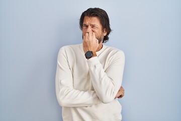Handsome middle age man wearing casual sweater over blue background looking stressed and nervous with hands on mouth biting nails. anxiety problem.