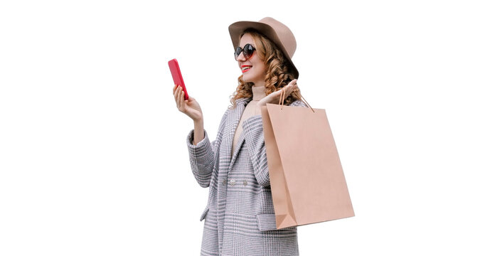 Portrait of happy smiling young woman taking selfie with smartphone holding shopping bags wearing coat, round hat isolated on white background