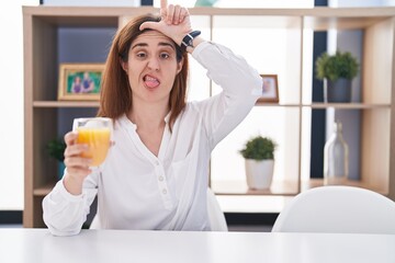 Brunette woman drinking glass of orange juice making fun of people with fingers on forehead doing loser gesture mocking and insulting.