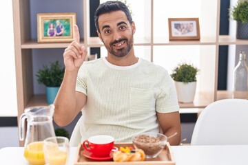 Hispanic man with beard eating breakfast smiling with an idea or question pointing finger up with happy face, number one