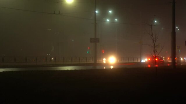 Cars with headlights on are moving along a city street at night in fog, rain. Silhouettes of cars, reflections on the pavement, blurry images.