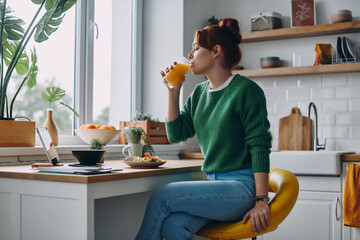 Beautiful redhead woman drinking juice while having lunch at the domestic kitchen