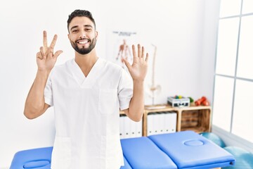 Young handsome man with beard working at pain recovery clinic showing and pointing up with fingers number eight while smiling confident and happy.