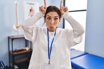 Young brunette woman working at pain recovery clinic doing funny gesture with finger over head as...