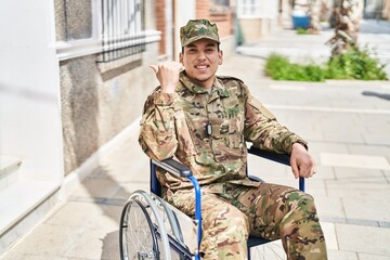 Young arab man wearing camouflage army uniform sitting on wheelchair pointing thumb up to the side smiling happy with open mouth