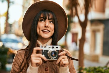 Brunette woman wearing winter hat smiling using vintage camera outdoors at the city - Powered by Adobe