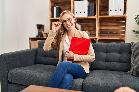 Young blonde woman psychologist smiling confident holding clipboard at psychology clinic
