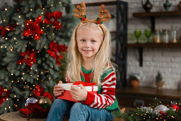 Merry Christmas and Happy Holidays. Surprised cute child girl opening present. Little kid having fun near Christmas tree indoors. Funny girl with gift box near christmas tree
