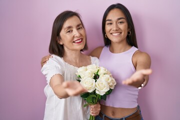 Hispanic mother and daughter holding bouquet of white flowers smiling cheerful offering palm hand...