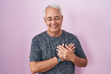 Middle age man with grey hair standing over pink background smiling with hands on chest with closed eyes and grateful gesture on face. health concept.