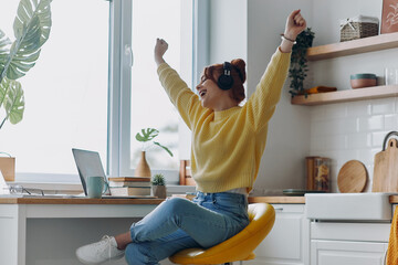 Happy woman in headphones keeping arms raised while sitting near the laptop at the kitchen counter