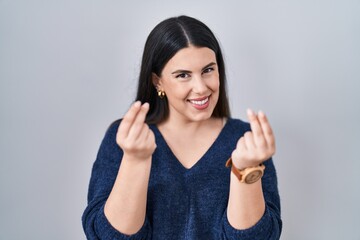 Young brunette woman standing over isolated background doing money gesture with hands, asking for salary payment, millionaire business