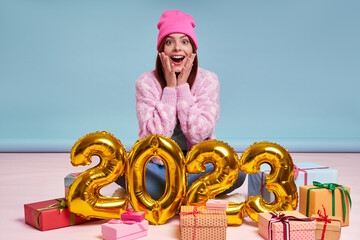 Surprised young woman sitting near the gift boxes and gold colored numbers against blue background