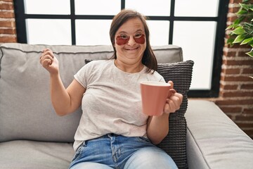 Hispanic girl with down syndrome sitting on the sofa drinking a coffee at home screaming proud, celebrating victory and success very excited with raised arm