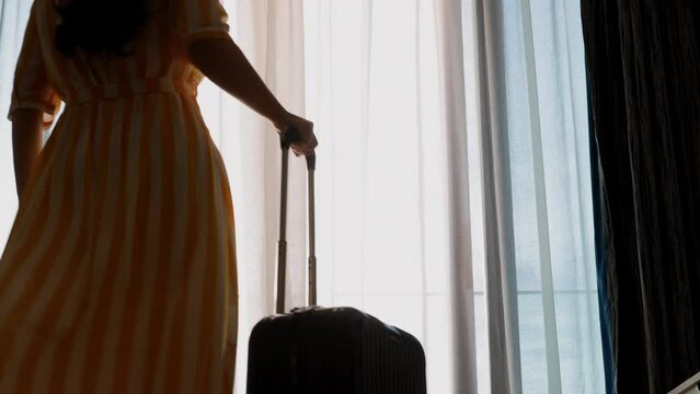 Asian businesswoman in yellow dress and yellow hat pulling her luggage in hotel bedroom, standing and enjoy watching the sea view at balcony. 
