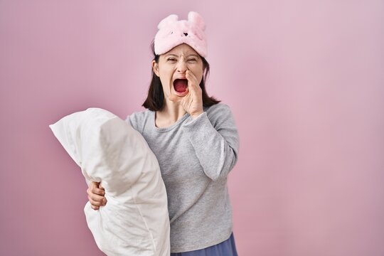 Woman with down syndrome wearing sleeping mask hugging pillow shouting and screaming loud to side with hand on mouth. communication concept.