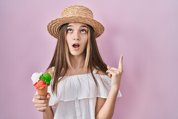 Teenager girl holding ice cream amazed and surprised looking up and pointing with fingers and raised arms.