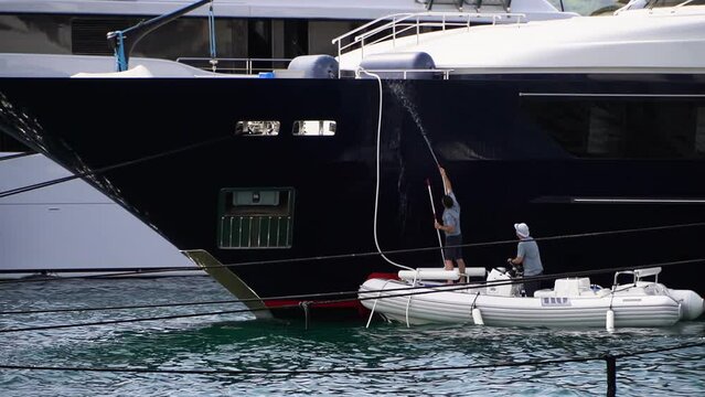 The crew of a huge boat washes a sailing yacht in clear sunny weather, the guys pour water from a hose and tinder with special mops, it is preparing the luxury boat