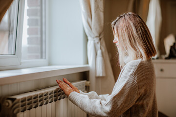 Young blond woman in long winter beige sweater is posing at home near the radiator. Winter season concept. Economy program 