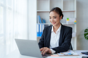 Happy Asian businesswoman sitting in the office with her laptop computer working diligently and smiling brightly with loads of papers on her desk.