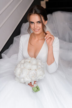 Portrait of elegant brunette bride in puffy white wedding dress with open neckline, sitting on stairs indoors holding beautiful bouquet of peonies. Holds a strand of hair and smiles.