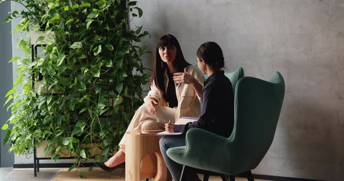 Older and younger multi ethnic colleagues talking eaten on armchairs in modern office lobby, intern listen to guidance of middle-aged woman mentor, give advice, provide helpful information. Interview