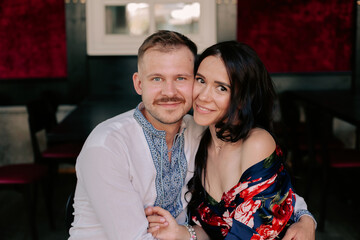 Fototapeta premium Portrait of caucasian couple man and woman embracing man looking at the camera. Happy man in shirt with blue embroidery embracing his attractive girlfriend with long hair, dressed in elegant dress, 