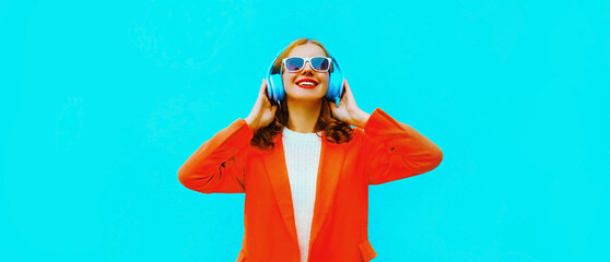 Portrait of happy smiling young woman in headphones listening to music wearing red jacket on blue background
