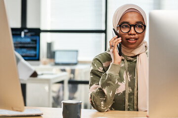 Young confident Muslim employee in hijab and eyeglasses consulting client on mobile phone while...