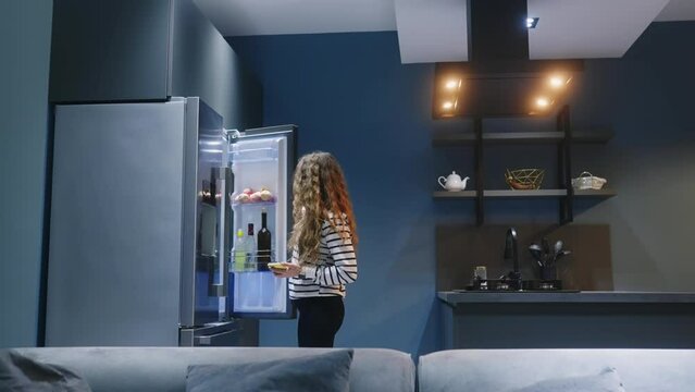 Woman enters kitchen, turns on lights with smartphone app in her smart home IoT system. Female uses application on phone to control lightning scenarios in living room, pouring wine. Internet of things