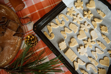 Top view. Selective focus on a baking sheet with carved molds of gingerbread dough, cookie cutters on floured wooden board and fir-tree needles and golden pine cones on the table. Christmas bakery