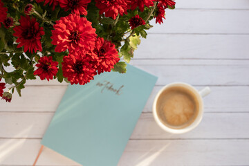 Red chrysanthemum flowers on blurred cup of coffee cappuccino and blue notebook on wooden table background. Top view, flat lay, copy space. Selective focus	