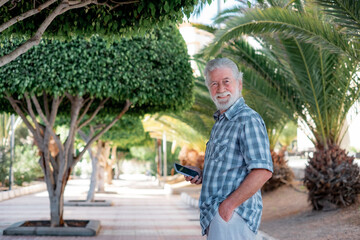 Attractive elderly man walks in the public park enjoying retirement and good weather. Senior bearded male smiles looking at camera holding mobile phone