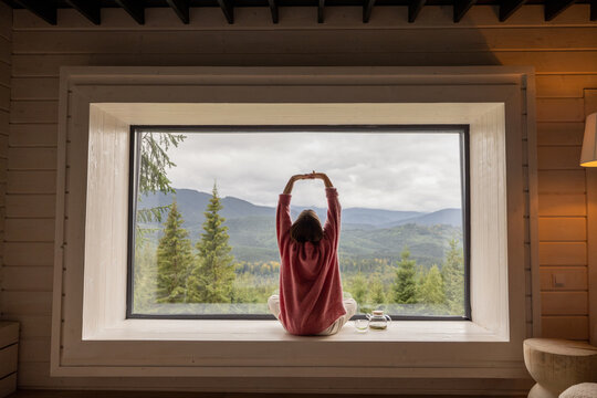 Woman meditates while sitting on window sill and enjoys panoramic view on mountains. Recreation and escaping to nature concept