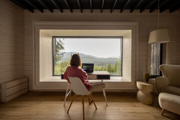 Woman works on laptop while sitting by the table in front of panoramic window with great view on...