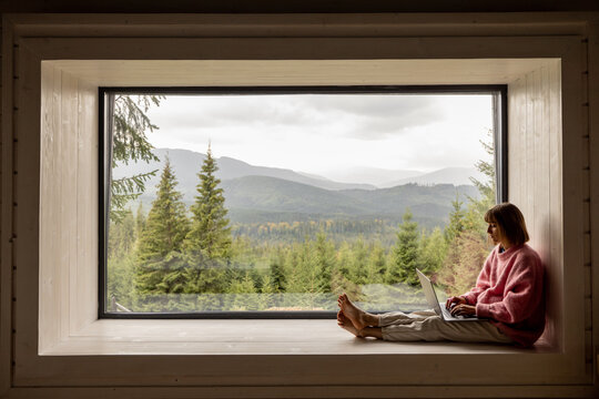 Fototapeta Woman works on laptop while sitting on a window with great view on mountains. Remote work from cozy place and escaping to nature concept