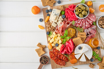 Charcuterie board with a variety of cheeses, salami, chorizzo, prosciutto, honey, grapes, nuts, olives, bread, blueberries and fresh herbs on a white wooden background. A festive snack.