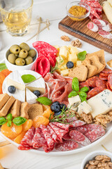 Charcuterie board with a variety of cheeses, salami, chorizzo, prosciutto, honey, grapes, nuts, olives, bread, blueberries and fresh herbs on a white wooden background. A festive snack.