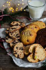 Mix of different kinds of Christmas cookies: biscotti, American chocolate cookies and nordic spicy gingerbread cookies  on baking paper and glass of milk on wooden table.