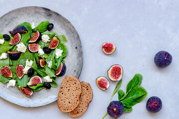 Fresh healthy summer salad with figs, white cheese, arugula and black olives on gray ceramic plate and two pieces of whole wheat bread on gray background. Top down view. Close-up.
